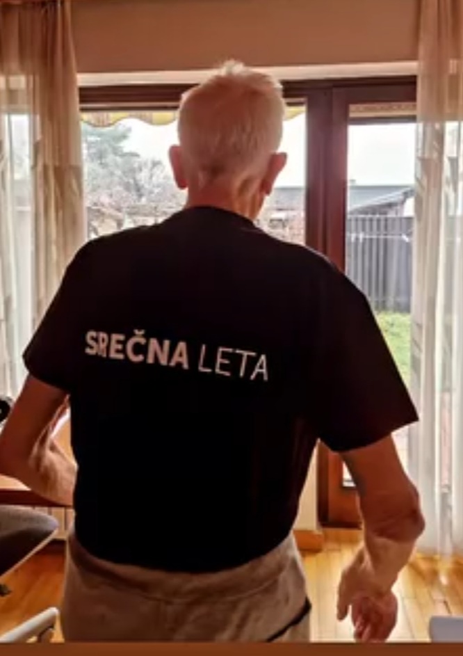 Starejša oseba stoji ob oknu, oblečena v majico z napisom "Srečna leta". Oskrbovalna ekipa podjetja Srečna leta ponuja kakovostno in sočutno oskrbo na domu za starejše v Ljubljani in okolici.