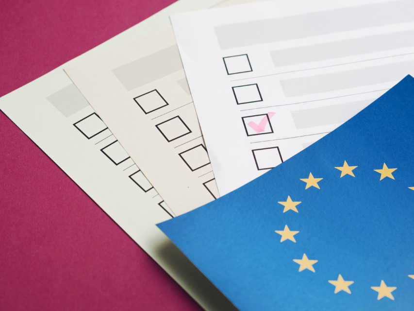 Na mizi so listi z obrazci in kljukicami, delno prekriti z zastavo Evropske unije.