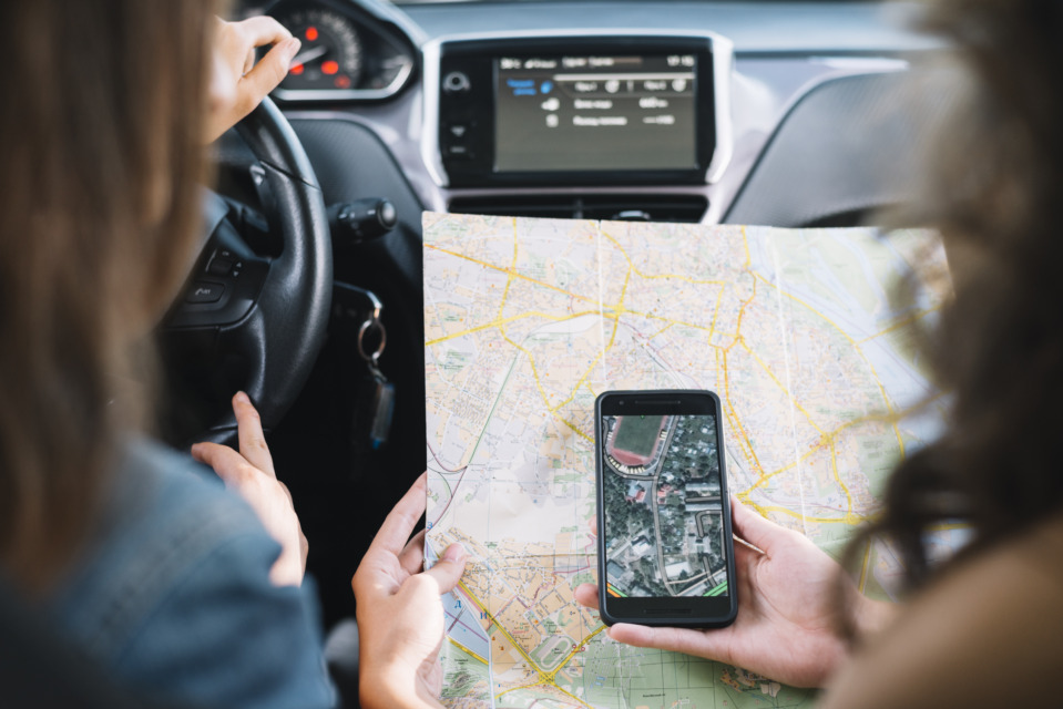 Dve osebi v avtomobilu pregledujeta papirni zemljevid in navigacijo na telefonu ter načrtujeta pot pred vožnjo.