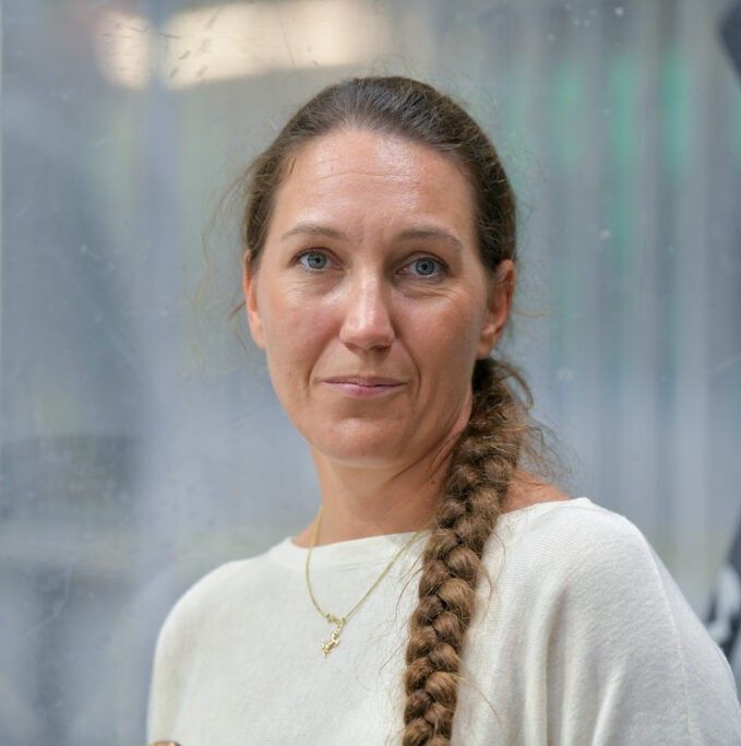 Portrait of a woman with a long braided hair over one shoulder, wearing a white sweater and a gold necklace, looking at the camera.