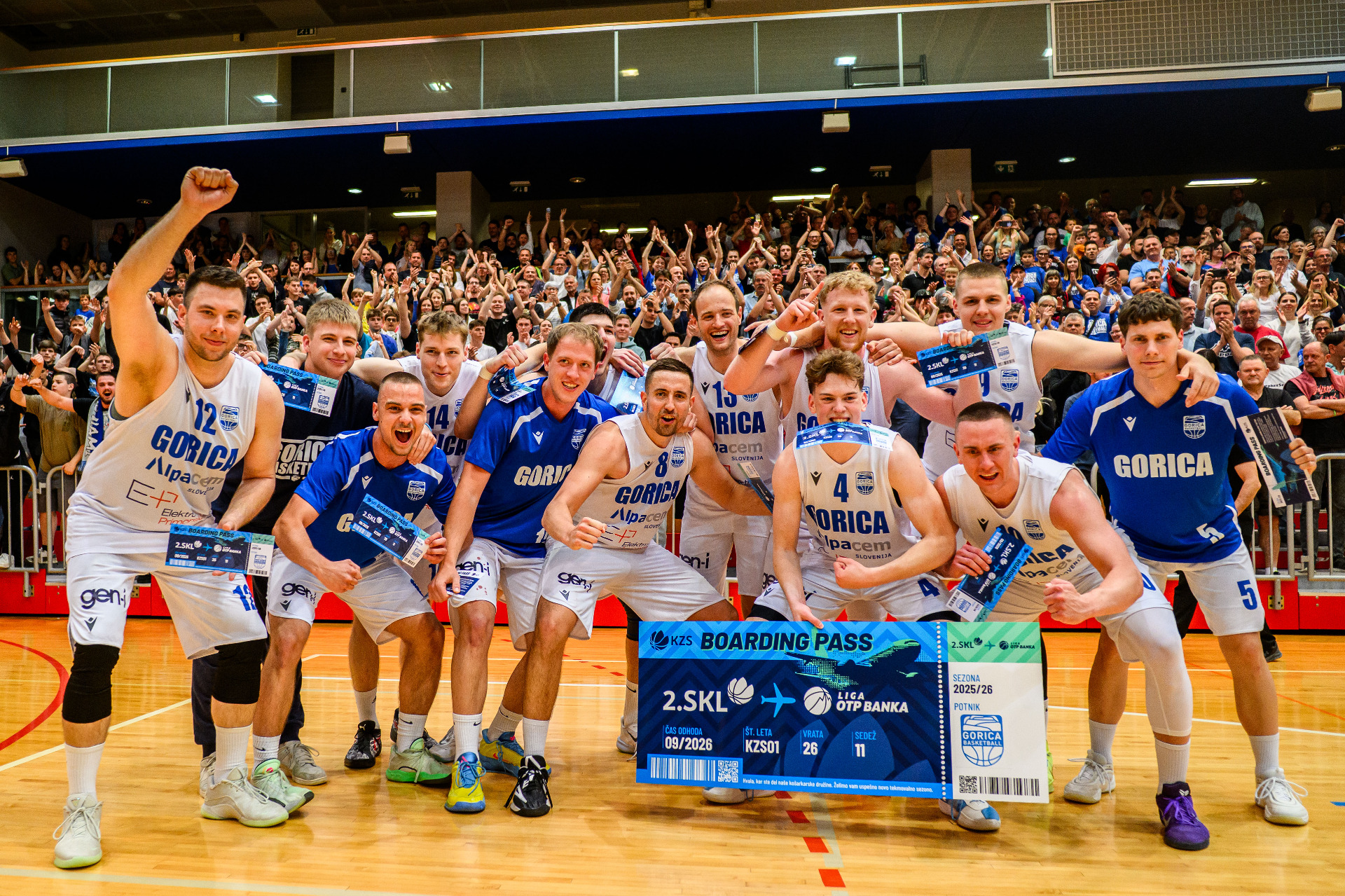Group of basketball players in white and blue jerseys pose with a cheering crowd in an indoor arena after a game.