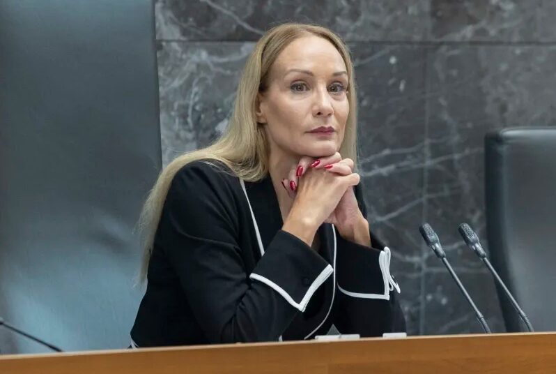 Woman in a black blazer with white trim sits at a wooden desk with two microphones, hands clasped under her chin, against a dark marble backdrop.