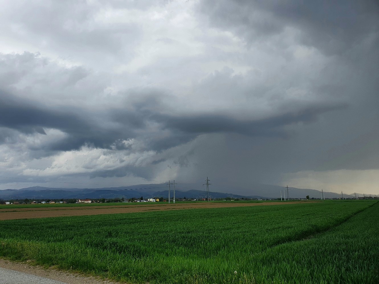 Dark storm clouds over green fields with power lines and distant hills; rain may be approaching.