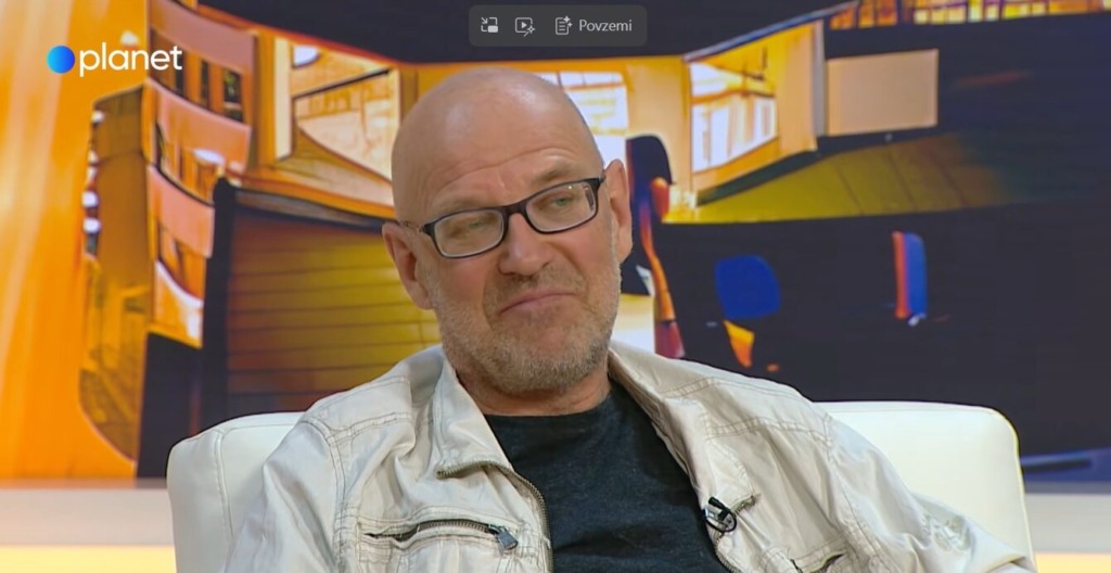 Guest on a talk-show set: bald man with glasses in a light jacket seated on a white chair, warm orange studio backdrop and planet logo.