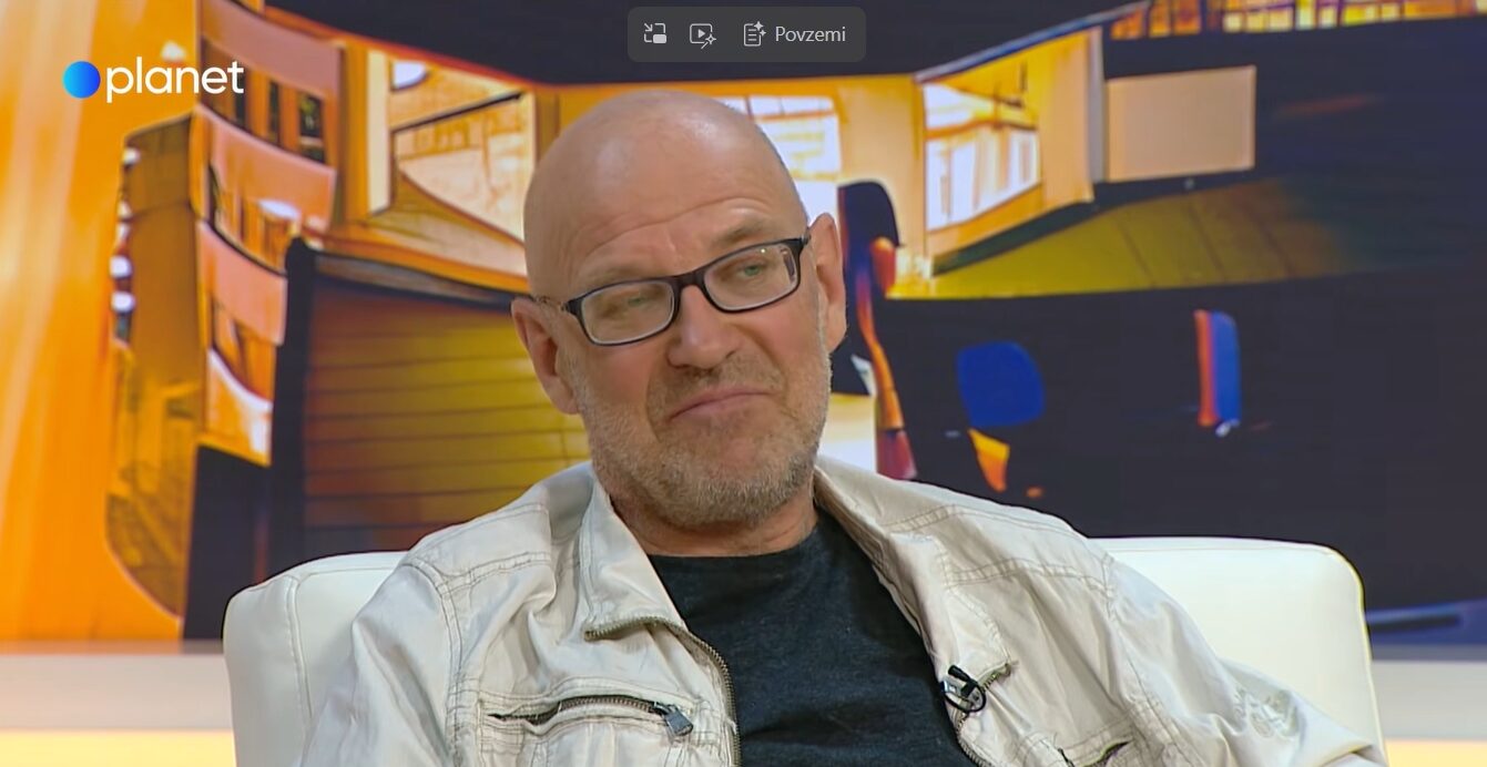 Guest on a talk-show set: bald man with glasses in a light jacket seated on a white chair, warm orange studio backdrop and planet logo.