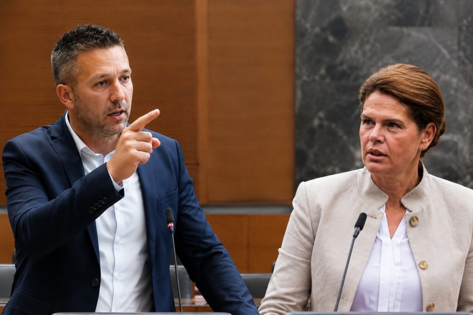 Man in a navy suit points while speaking at a formal meeting, with a woman listening beside him and microphones in front.