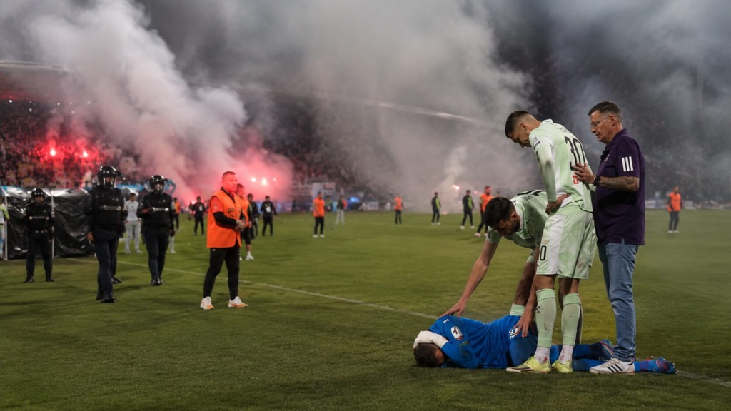 Soccer players surround a fallen goalkeeper on the field while smoke flares glow in the stands and fans cheer in the background.