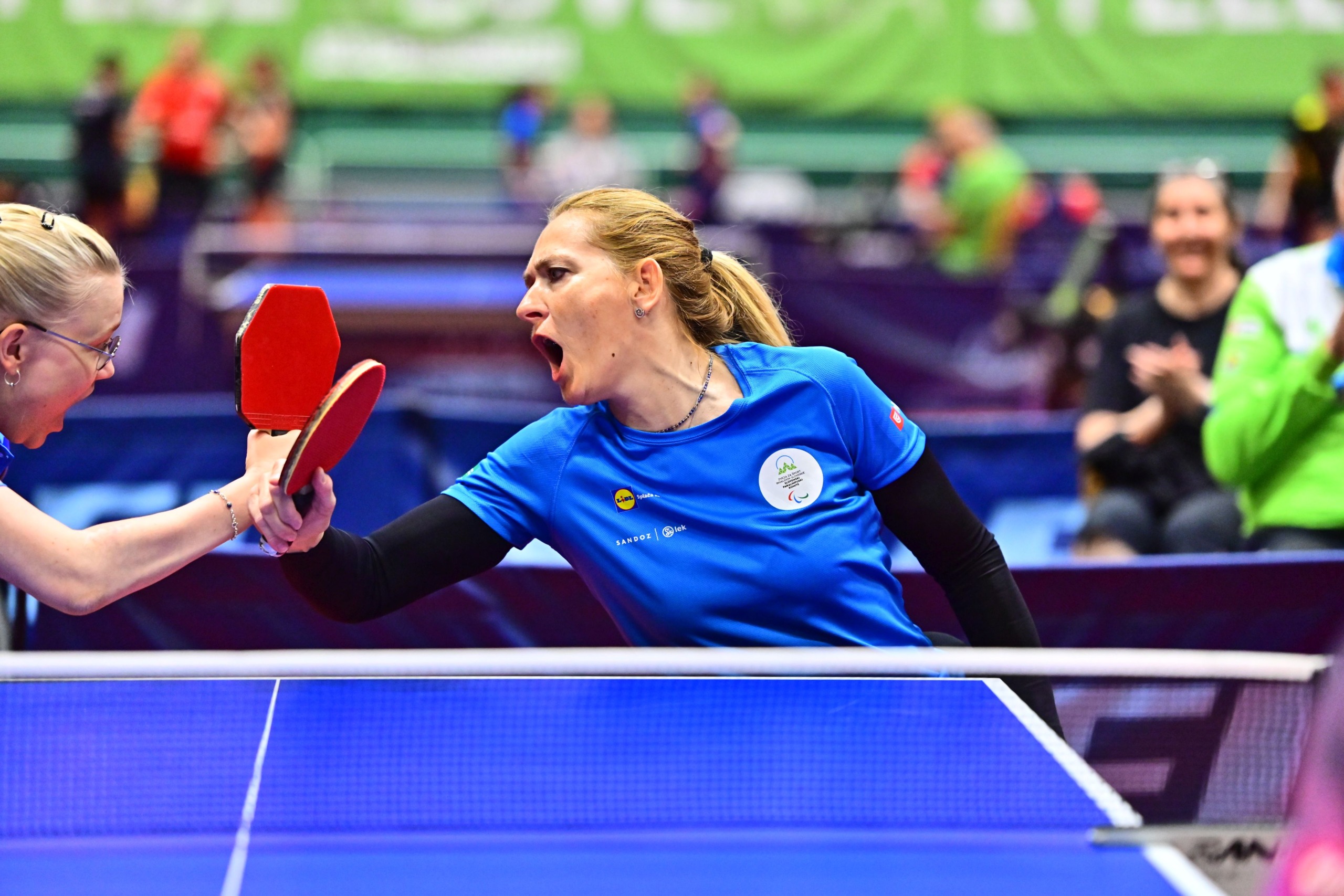 Two women playing table tennis in blue shirts clash at the net during a rally.