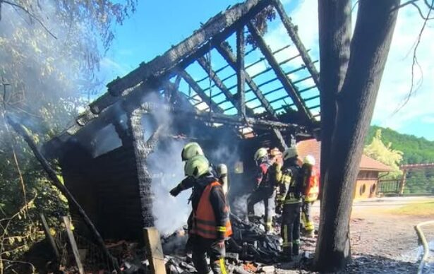 Firefighters in full gear extinguish a partially burned wooden building, smoke rising from the charred roof and beams.