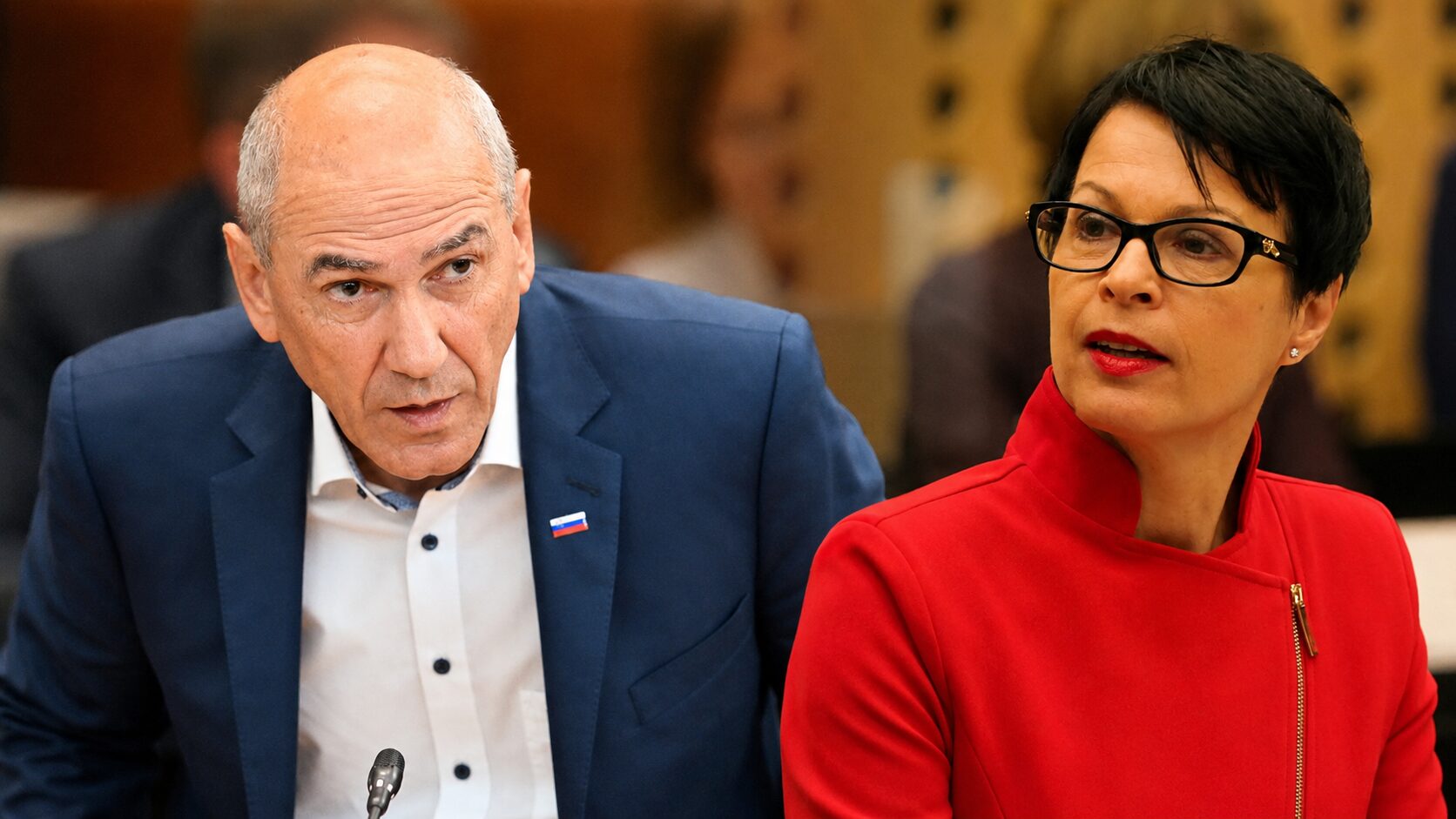Two individuals seated at a table in a formal setting; man in blue suit glances toward the woman, who wears a red outfit and glasses and looks to the side.