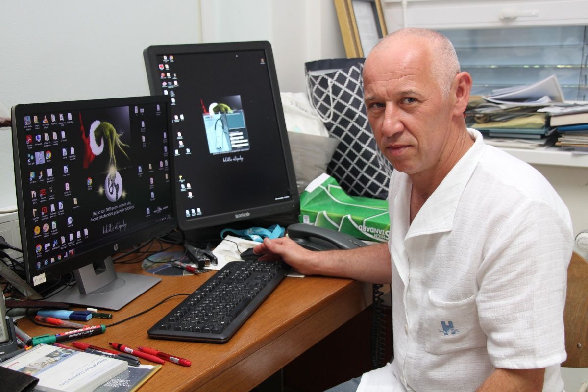 Man in a white short-sleeve shirt sits at a cluttered desk with two computer monitors and a keyboard, looking at the camera.