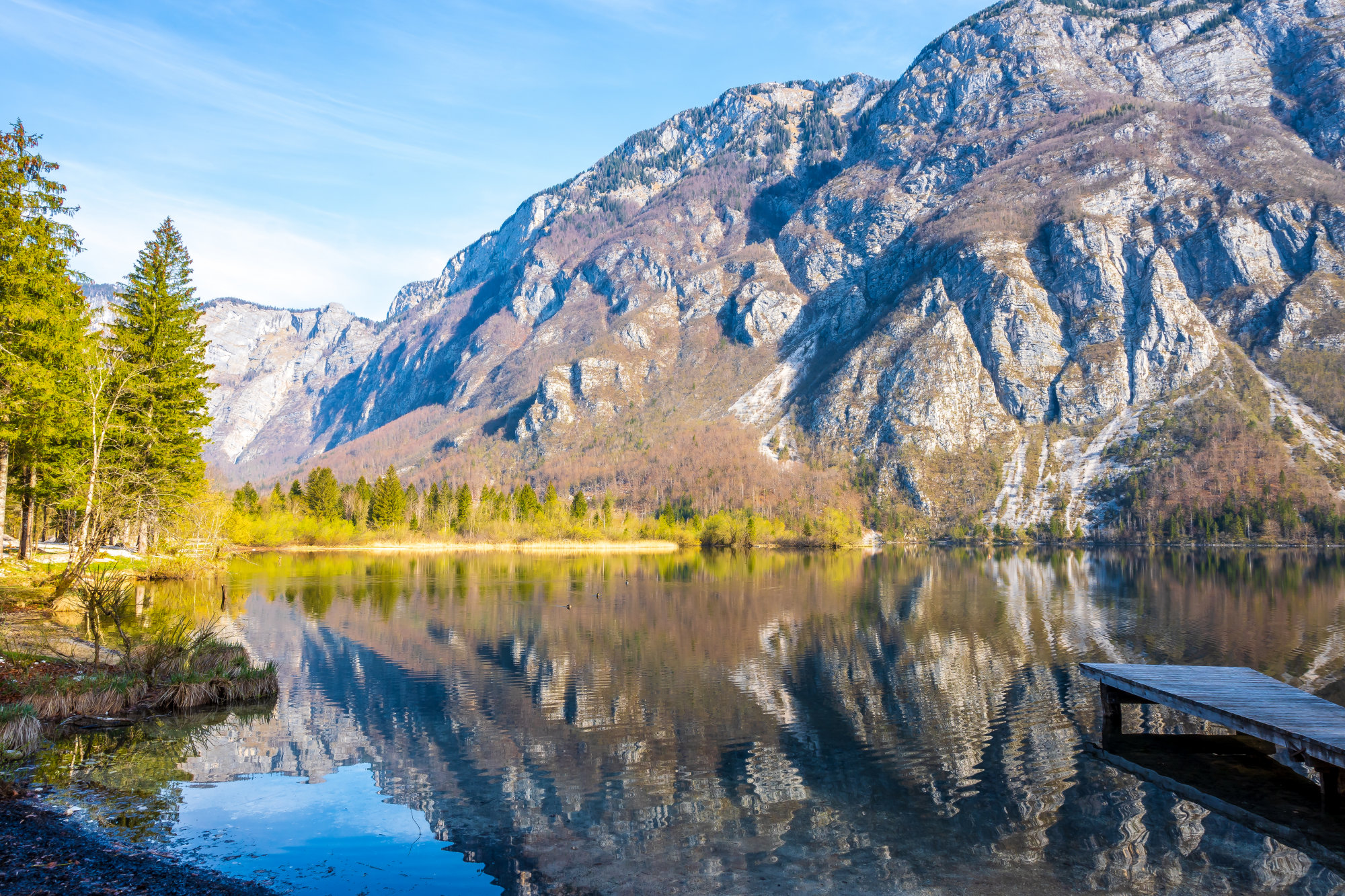 Mountain range with rocky cliffs reflected in a calm lake; pine trees line the shore and a wooden dock appears on the right edge.
