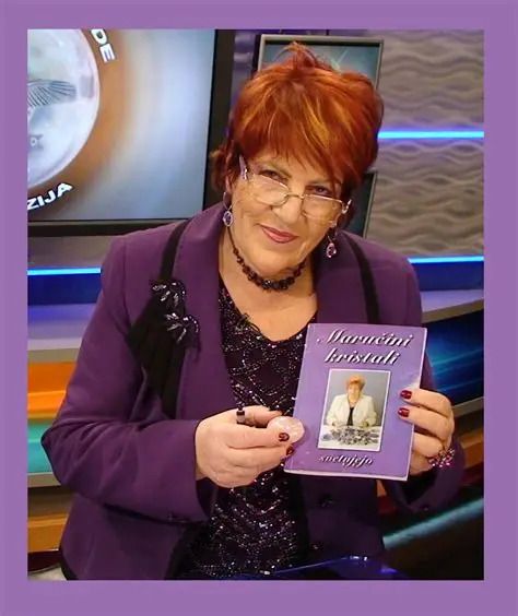 TV host in a purple outfit holding a small book with a photo on the cover, seated in a studio set with blue accents and a purple border behind her.