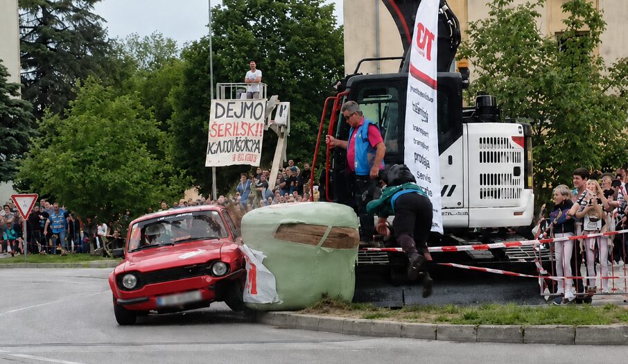 Red vintage car collides with a foam obstacle as a crowd watches a street stunt event near a large construction vehicle on the curbside search area?