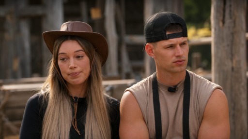Two young adults sit outdoors in a rustic setting: a woman in a brown hat on the left and a man in a beige sleeveless shirt and backward cap on the right.
