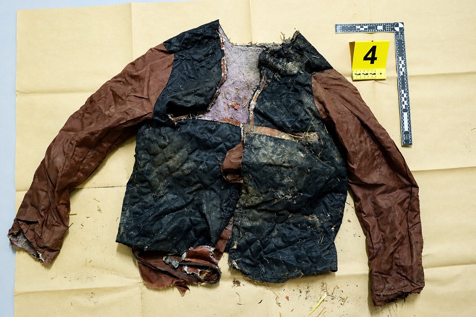 Torn quilted jacket with brown sleeves and a black body, dirty and damaged, laid on a beige surface next to a measuring ruler and a yellow evidence marker labeled 4.