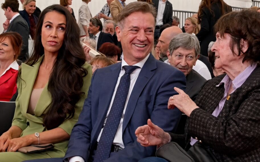 Smiling man in a blue suit conversation with an older woman at a formal event, with a woman in a green blazer seated beside him.