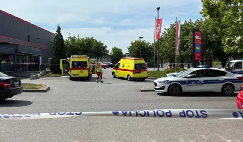Emergency scene outside a building with yellow ambulances, police car, and responders behind police tape marked Stop Policja/Area Secured.