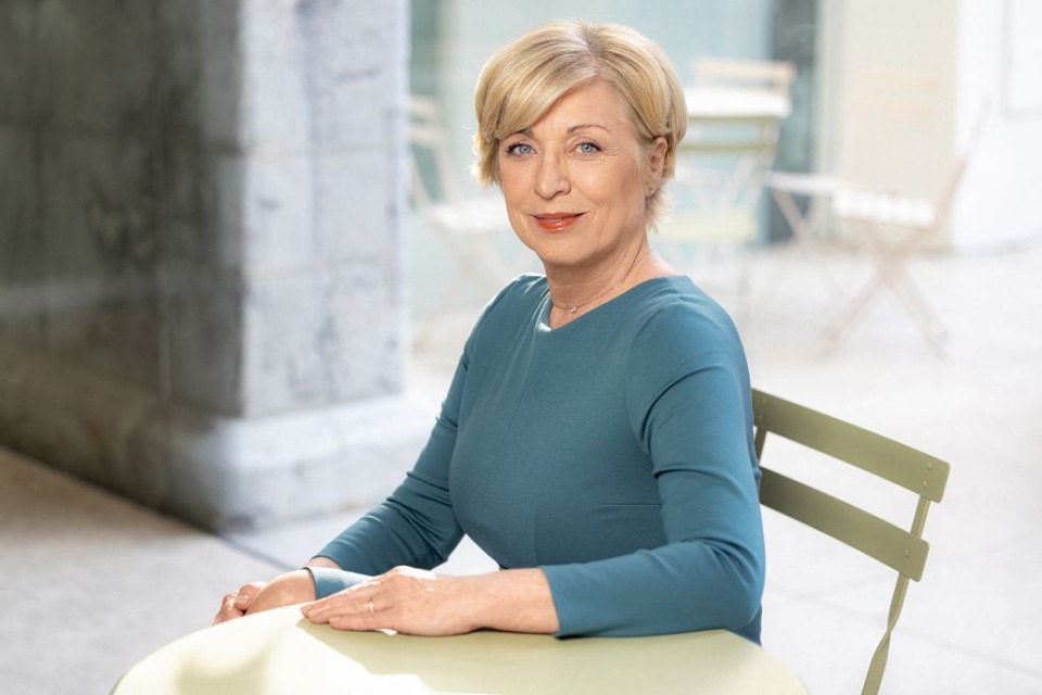 Smiling woman with blonde hair seated at an outdoor cafe table in a teal top.