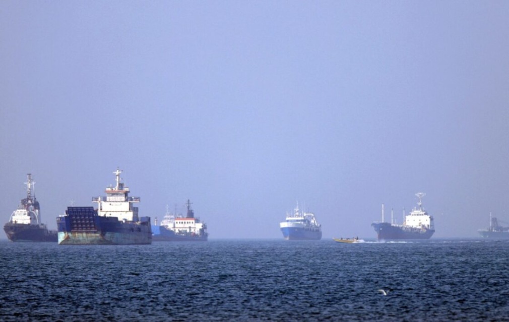 Several large cargo ships sit anchored on a calm blue sea near the horizon with a clear sky above.