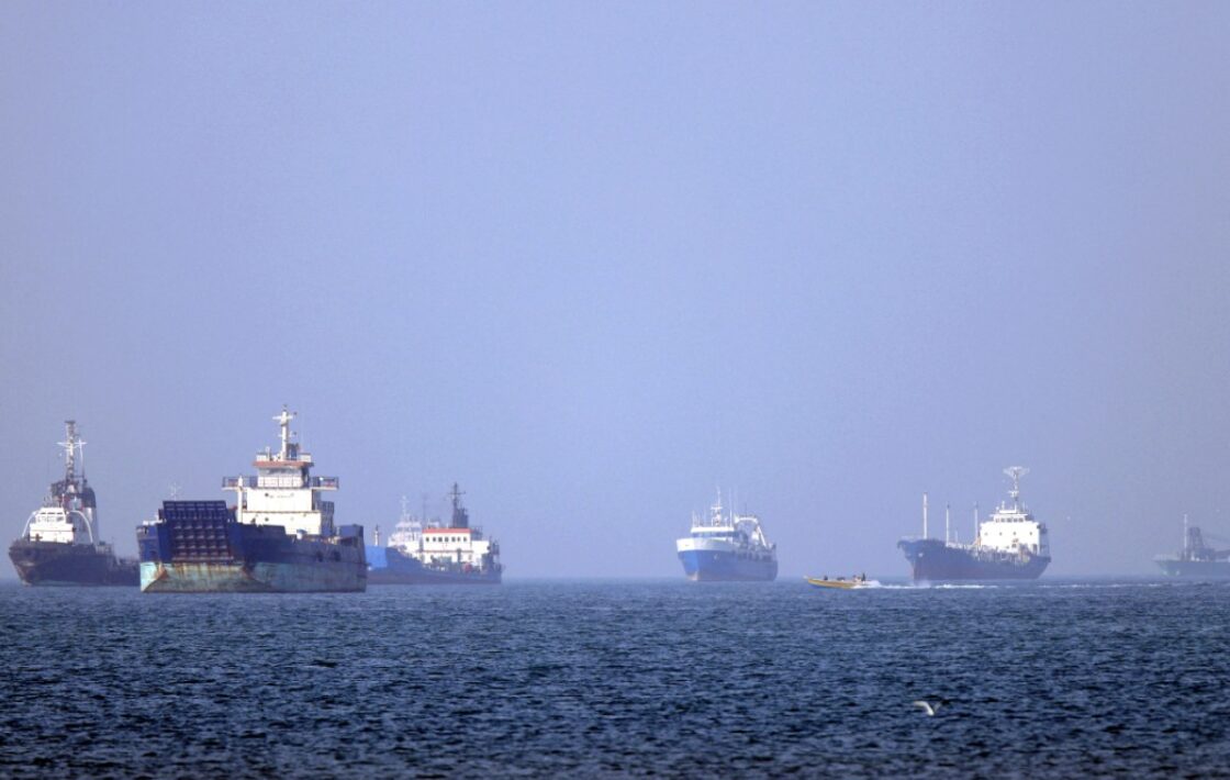 Several large cargo ships sit anchored on a calm blue sea near the horizon with a clear sky above.