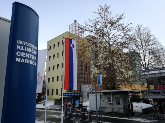 Tall blue sign reading 'Univerzitetni klinični center Maribor' beside a hospital building with a vertical Slovenian flag in winter light.