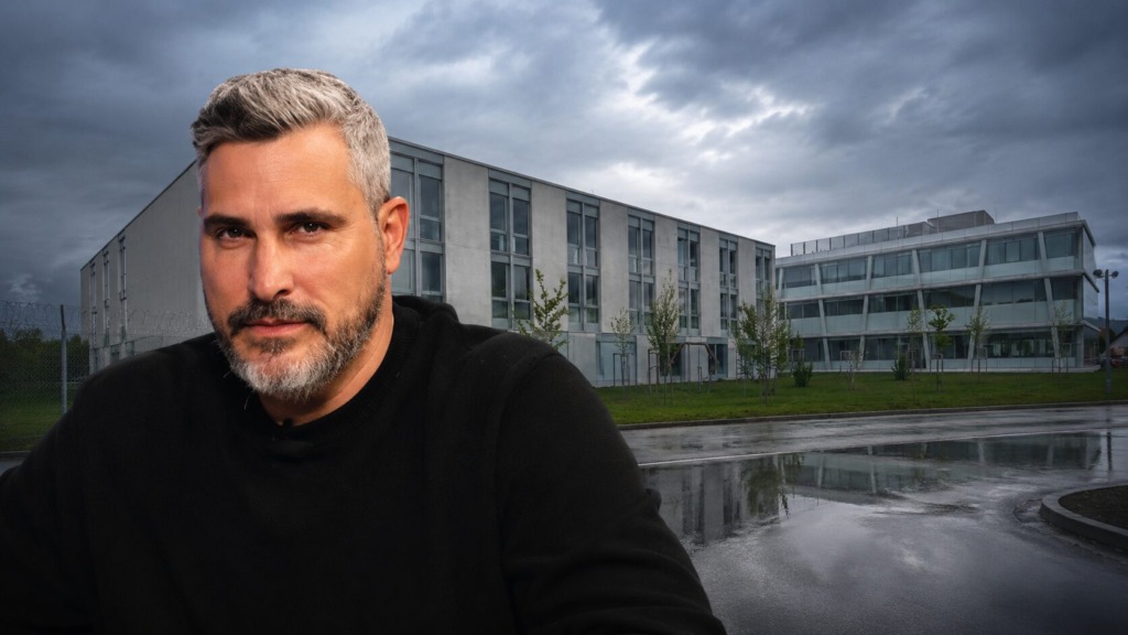 Portrait of a middle-aged man with gray hair and a beard in a black sweater, outdoors beside a modern office complex on a cloudy day.