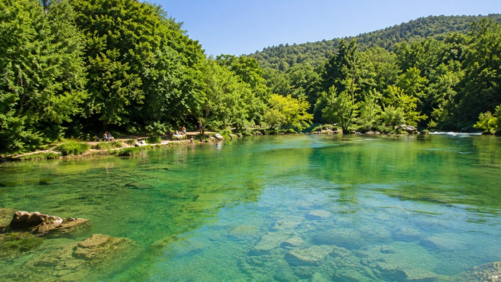 Mirna in čista reka Krka z zelenimi gozdnatimi bregovi ter naravnim prostorom za kopanje.